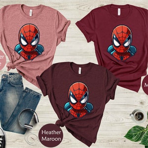 May include: Three t-shirts in heather mauve, maroon, and heather maroon, each featuring a cartoon Spider-Man graphic. The shirts are displayed with blue jeans, headphones, and sneakers. The text "Heather Mauve", "Maroon", and "Heather Maroon" are visible.