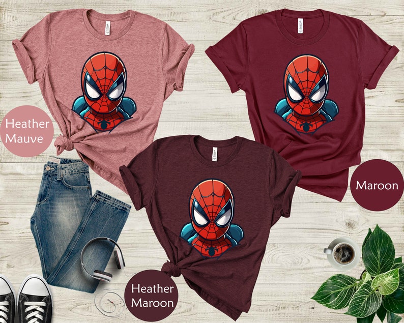 May include: Three t-shirts in heather mauve, maroon, and heather maroon, each featuring a cartoon Spider-Man graphic. The shirts are displayed with blue jeans, headphones, and sneakers. The text "Heather Mauve", "Maroon", and "Heather Maroon" are visible.