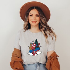 Puede incluir: Camiseta gris claro con un dise&ntilde;o de Spider-Man navide&ntilde;o. La ilustraci&oacute;n muestra a Spider-Man con un gorro de Pap&aacute; Noel, arrodillado junto a un &aacute;rbol de Navidad y un saco de regalos. El dise&ntilde;o es de estilo caricatura.