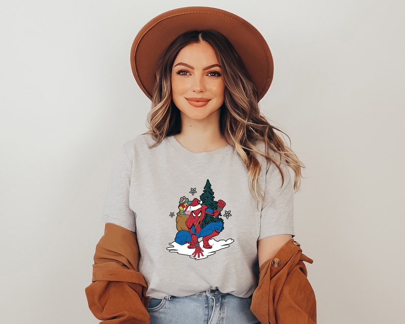 Puede incluir: Camiseta gris claro con un dise&ntilde;o de Spider-Man navide&ntilde;o. La ilustraci&oacute;n muestra a Spider-Man con un gorro de Pap&aacute; Noel, arrodillado junto a un &aacute;rbol de Navidad y un saco de regalos. El dise&ntilde;o es de estilo caricatura.