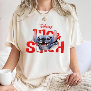 Puede incluir: Camiseta color crema con el logo de Disney y las palabras "Lilo & Stitch" en rojo. El gr&aacute;fico muestra un personaje alien&iacute;gena azul sonriendo que rompe la camiseta. Una camiseta casual y c&oacute;moda.