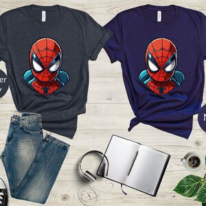 May include: Two t-shirts, one heather navy and one navy, each featuring a cartoon Spider-Man graphic. The superhero's face is red with white eyes and a black spider emblem. The shirts are paired with jeans, sneakers, a notebook, and headphones.