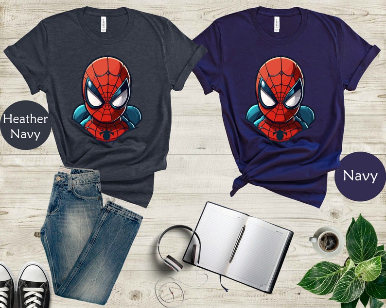 May include: Two t-shirts, one heather navy and one navy, each featuring a cartoon Spider-Man graphic. The superhero's face is red with white eyes and a black spider emblem. The shirts are paired with jeans, sneakers, a notebook, and headphones.
