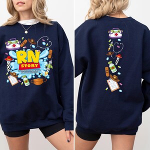 K&ouml;nnte beinhalten: Marineblaues Sweatshirt mit farbenfrohem Grafikdesign. Auf der Vorderseite steht "RN STORY" mit medizinischen und Cowboy-Illustrationen. Die R&uuml;ckseite des Sweatshirts hat eine vertikale Anordnung &auml;hnlicher Illustrationen. Der Satz "TO INFINITE AND BEYOND FOR PATIENTS" ist ebenfalls sichtbar.