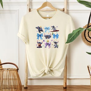 Puede incluir: Camiseta color crema con un gr&aacute;fico colorido del personaje Stitch de Lilo & Stitch. El dise&ntilde;o incluye a Stitch en varias poses con lazos azules y morados. La camiseta se muestra en una percha de madera.
