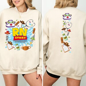 K&ouml;nnte beinhalten: Beiges Sweatshirt mit farbenfrohem Grafikdesign. Auf der Vorderseite steht "RN STORY" mit medizinischen und Cowboy-Illustrationen. Die R&uuml;ckseite hat eine vertikale Anordnung &auml;hnlicher Illustrationen. Das Sweatshirt besteht aus einem weichen Material.
