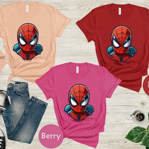 May include: Three t-shirts in heather peach, red, and berry colors, each featuring a cartoon Spider-Man graphic. The image includes a pair of blue jeans, black and white sneakers, and a pair of headphones.