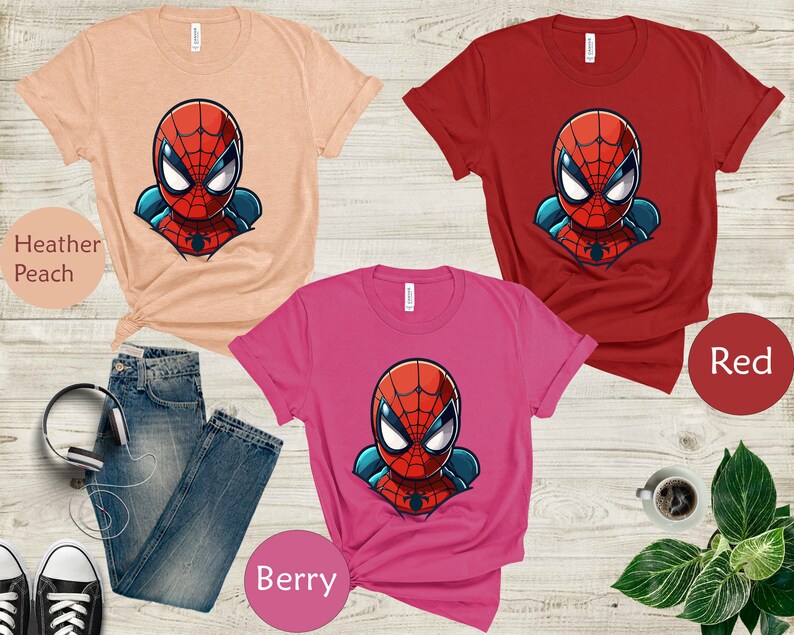 May include: Three t-shirts in heather peach, red, and berry colors, each featuring a cartoon Spider-Man graphic. The image includes a pair of blue jeans, black and white sneakers, and a pair of headphones.
