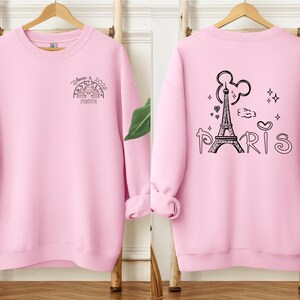 May include: A light pink sweatshirt with black graphic designs. The front features a castle design with the text "Disney 2024 Mom". The back displays the Eiffel Tower, Mickey Mouse ears, and the word "Paris".