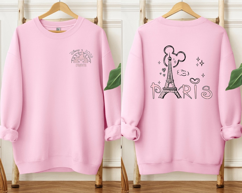 May include: A light pink sweatshirt with black graphic designs. The front features a castle design with the text "Disney 2024 Mom". The back displays the Eiffel Tower, Mickey Mouse ears, and the word "Paris".