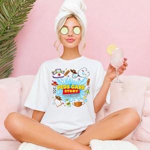 May include: White t-shirt with a colorful graphic design featuring medical-themed illustrations and the text "Peds Care Story". The design includes a stethoscope, cowboy hat, rocket, and other medical symbols. The shirt is worn by a person with a towel on their head.