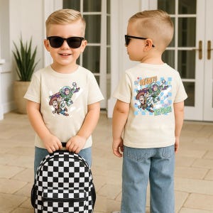 Puede incluir: Un ni&ntilde;o lleva una camiseta color crema con Buzz Lightyear y Woody de Toy Story. La camiseta tiene la frase "&iexcl;Hasta el infinito y m&aacute;s all&aacute;!" en la espalda. El ni&ntilde;o lleva vaqueros azules y una mochila a cuadros blancos y negros.