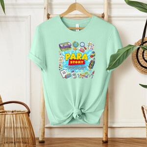 K&ouml;nnte beinhalten: Hellgr&uuml;nes T-Shirt mit dem Schriftzug "Para Story" in Gelb und Rot, umgeben von farbenfrohen Illustrationen von Schulmaterialien und Spielzeug. Das T-Shirt hat einen Rundhalsausschnitt und kurze &Auml;rmel und h&auml;ngt an einem Holzb&uuml;gel.