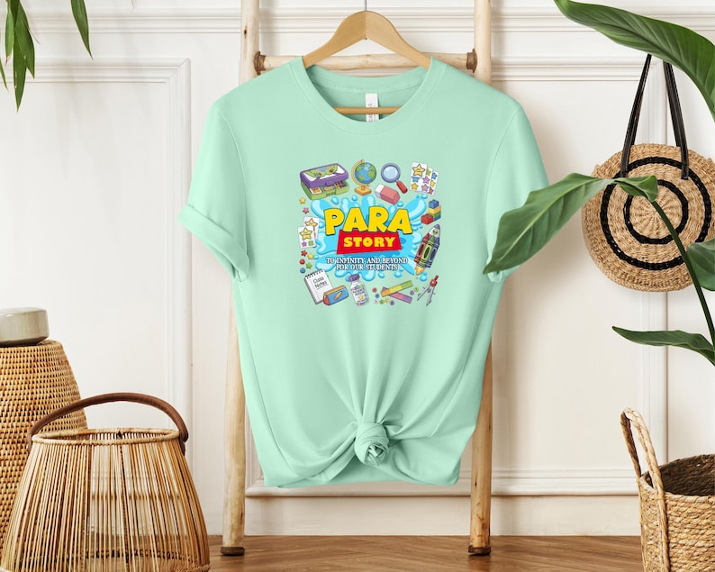 K&ouml;nnte beinhalten: Hellgr&uuml;nes T-Shirt mit dem Schriftzug "Para Story" in Gelb und Rot, umgeben von farbenfrohen Illustrationen von Schulmaterialien und Spielzeug. Das T-Shirt hat einen Rundhalsausschnitt und kurze &Auml;rmel und h&auml;ngt an einem Holzb&uuml;gel.
