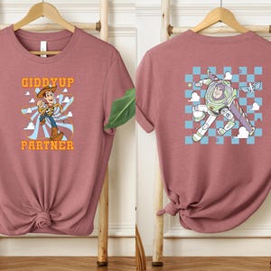 May include: Dusty rose t-shirt featuring a cartoon Woody character with the text "Giddyup Partner" on the front. The back of the shirt displays a cartoon Buzz Lightyear character. The shirt is displayed on a wooden rack.
