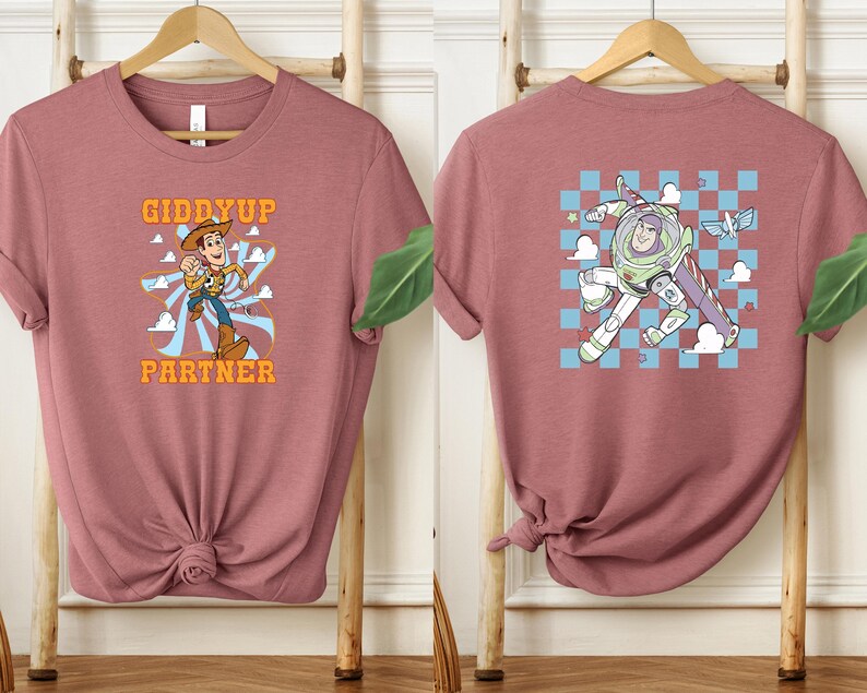 May include: Dusty rose t-shirt featuring a cartoon Woody character with the text "Giddyup Partner" on the front. The back of the shirt displays a cartoon Buzz Lightyear character. The shirt is displayed on a wooden rack.
