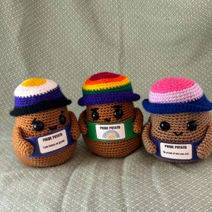 May include: Three brown crocheted potato figures with black eyes and small smiles. Each wears a different colored hat and holds a sign. The signs read: PRIDE POTATO Love knows no gender, PRIDE POTATO with a rainbow, and PRIDE POTATO Be proud of who you are.