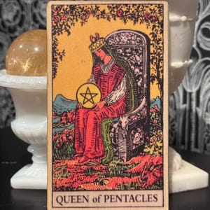 May include: A tarot card featuring the Queen of Pentacles. The card depicts a woman in a red gown and crown, holding a golden pentacle. The background shows a garden scene with a throne. The card's title, "QUEEN of PENTACLES," is at the bottom.