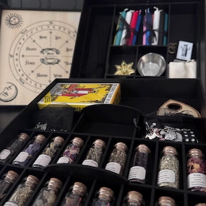 May include: A black wooden box filled with various spiritual items. The box contains small glass bottles with herbs, candles, tarot cards, a metal bowl, and a wooden planchette. A wooden board with a sun and moon design is visible in the background.