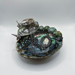 May include: An iridescent abalone shell serving as a jewelry dish, adorned with a silver necklace and decorative stones. The shell's interior displays a spectrum of colors, including blues, greens, and purples, creating a unique display for small items.