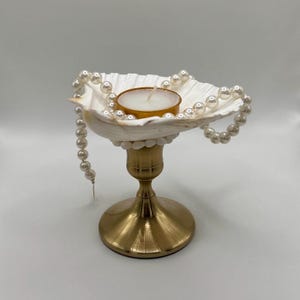 Op de afbeelding: Een decoratieve kandelaar met een witte schelpenkom, versierd met een parelketting. De kom bevat een kleine kaars. De houder heeft een goudkleurige basis, wat een elegante en kustge&iuml;nspireerde esthetiek cre&euml;ert.