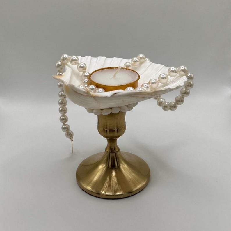 Op de afbeelding: Een decoratieve kandelaar met een witte schelpenkom, versierd met een parelketting. De kom bevat een kleine kaars. De houder heeft een goudkleurige basis, wat een elegante en kustge&iuml;nspireerde esthetiek cre&euml;ert.