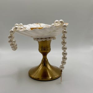 Op de afbeelding: Een decoratieve schelpvormige schaal met een goudkleurige metalen basis. De schaal is gevuld met kleine items en versierd met een parelsnoer. De parels vallen langs de zijkant naar beneden en geven de display een elegante uitstraling.