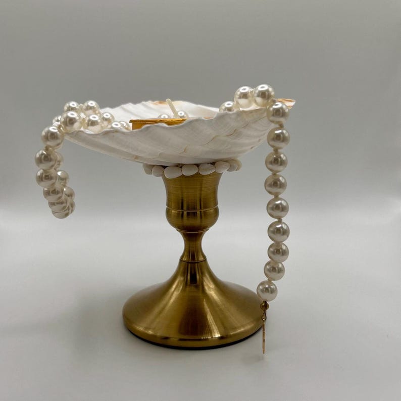 Op de afbeelding: Een decoratieve schelpvormige schaal met een goudkleurige metalen basis. De schaal is gevuld met kleine items en versierd met een parelsnoer. De parels vallen langs de zijkant naar beneden en geven de display een elegante uitstraling.