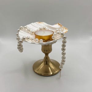 May include: A decorative candle holder featuring a white seashell, a gold-colored candle, and a strand of faux pearls. The candle holder has a gold-colored base. The candle is in a gold-colored metal container.