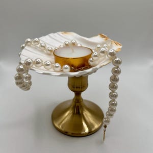 Op de afbeelding: Een decoratieve kandelaar met een schelpvormige schaal, een goudkleurige basis en een witte kaars in een goudkleurige houder. Een streng imitatieparels is om de zeeschelp gedrapeerd. De kandelaar is een decoratief item.