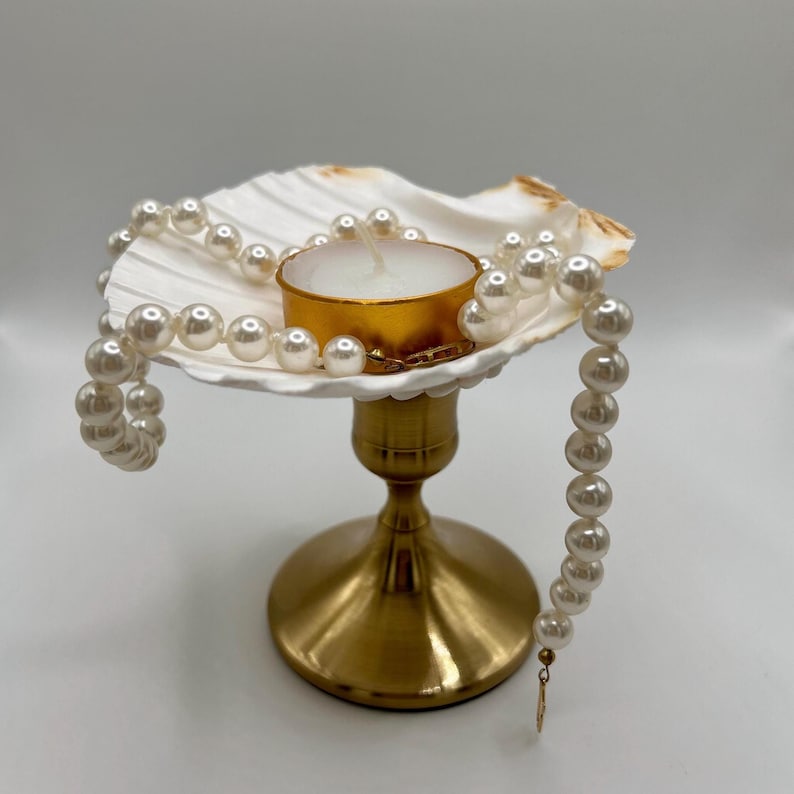 Op de afbeelding: Een decoratieve kandelaar met een schelpvormige schaal, een goudkleurige basis en een witte kaars in een goudkleurige houder. Een streng imitatieparels is om de zeeschelp gedrapeerd. De kandelaar is een decoratief item.