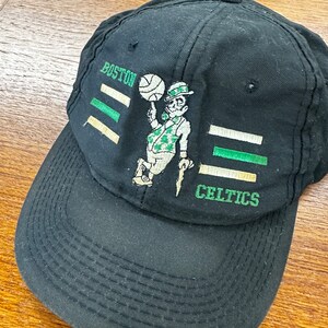 May include: Black baseball cap with a green and white embroidered logo of a cartoon leprechaun holding a basketball. The logo says "Boston Celtics".