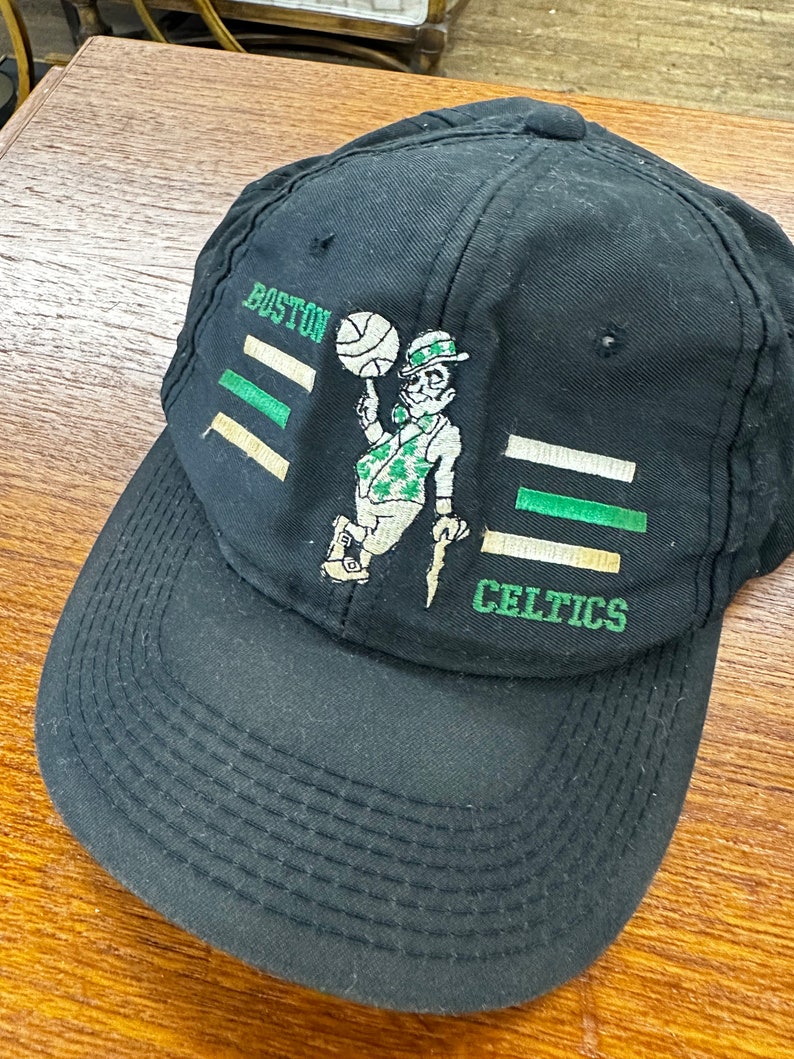 May include: Black baseball cap with a green and white embroidered logo of a cartoon leprechaun holding a basketball. The logo says "Boston Celtics".