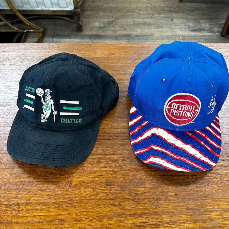 May include: Two baseball caps, one black with a green Boston Celtics logo and the other blue with a red Detroit Pistons logo.