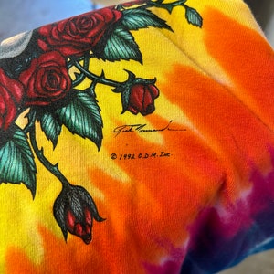 Puede incluir: Una camiseta tie-dye con un dise&ntilde;o colorido que presenta rosas y una firma. El texto "&copy; 1992 G.D.M. Inc." est&aacute; impreso en la camiseta.