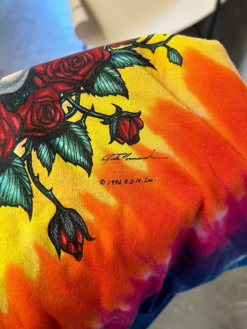 Puede incluir: Una camiseta tie-dye con un dise&ntilde;o colorido que presenta rosas y una firma. El texto "&copy; 1992 G.D.M. Inc." est&aacute; impreso en la camiseta.