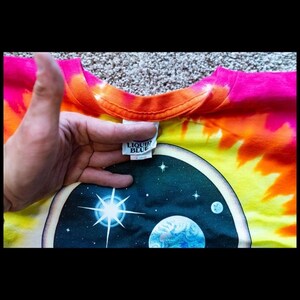 Puede incluir: Una camiseta tie-dye con un gr&aacute;fico de escena espacial. La camiseta es naranja, amarilla, rosa y azul. El gr&aacute;fico presenta un planeta, una estrella y una luna. La camiseta tiene una etiqueta Liquid Blue.
