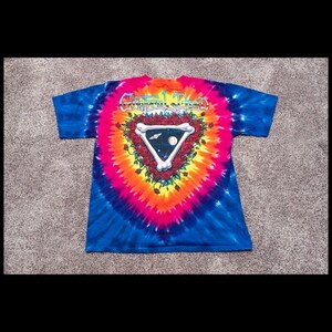 Puede incluir: Una camiseta tie-dye con un logotipo de Grateful Dead que presenta una calavera y rosas. El logotipo tiene forma de tri&aacute;ngulo con las palabras "Grateful Dead" encima.