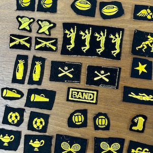 May include: A collection of 25 black felt patches with yellow embroidered designs. The patches feature various sports and activities, including baseball, football, tennis, swimming, band, and more.