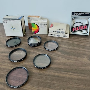May include: A collection of circular camera lens filters with black metal frames and their original cardboard packaging boxes. The boxes feature brand names including Hoya, Tiffen, and various filter specifications like 49mm, 52mm, and 55mm.