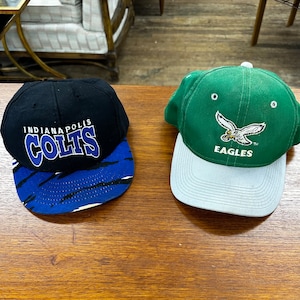May include: Two baseball caps, one black with blue and white stripes and the words "Indianapolis Colts" on the front, and one green with a white eagle and the words "Eagles" on the front.