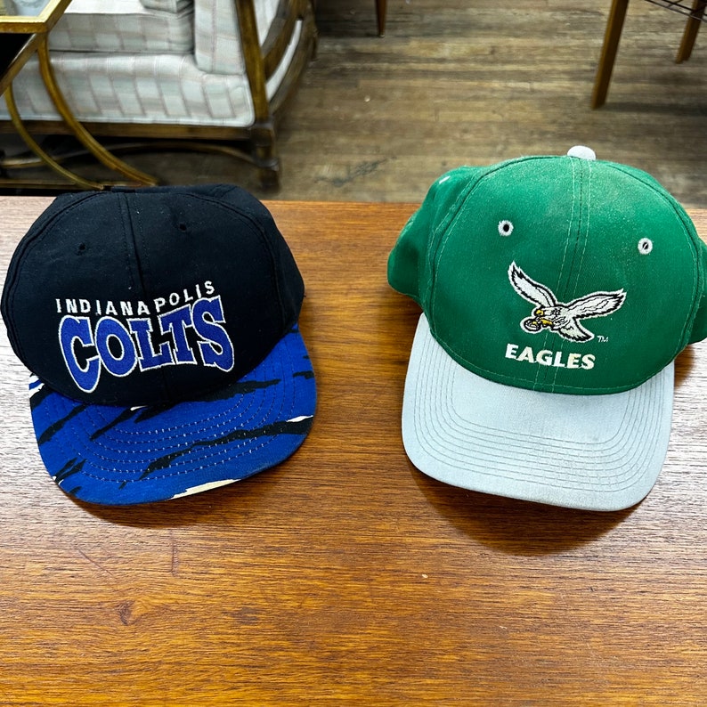 May include: Two baseball caps, one black with blue and white stripes and the words "Indianapolis Colts" on the front, and one green with a white eagle and the words "Eagles" on the front.