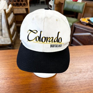 May include: White baseball cap with black brim. The front of the cap has the words "Colorado Buffaloes" embroidered in black and gold.