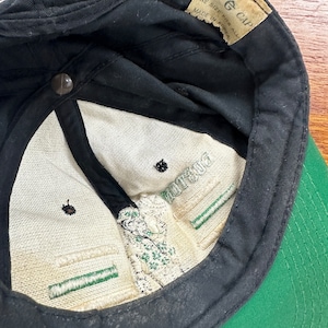 May include: Black and green baseball cap with a white label that reads "The G Cap" and "One Size Fits All Made in Korea".