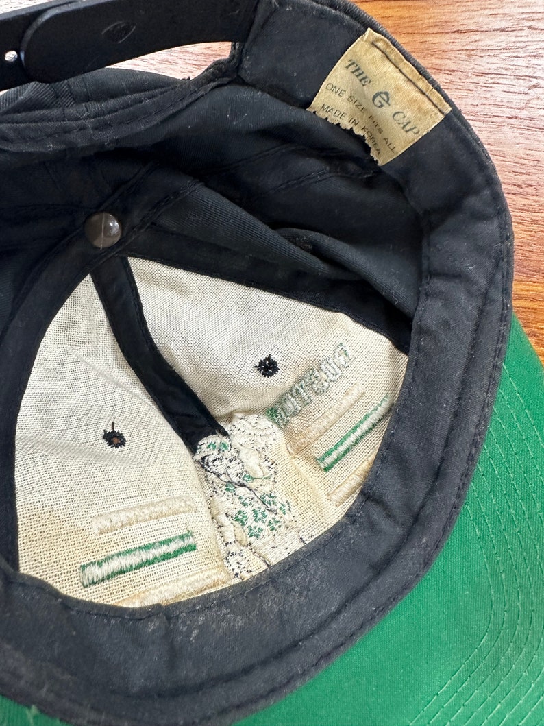 May include: Black and green baseball cap with a white label that reads "The G Cap" and "One Size Fits All Made in Korea".