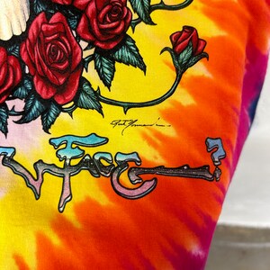 Puede incluir: Una camiseta tie-dye con un gr&aacute;fico colorido de una calavera con rosas y el texto "Face". La camiseta tiene un patr&oacute;n tie-dye amarillo, naranja, rosa y azul.