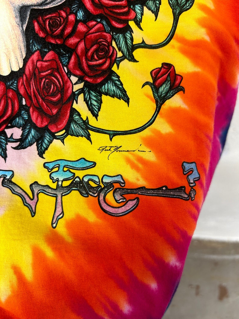 Puede incluir: Una camiseta tie-dye con un gr&aacute;fico colorido de una calavera con rosas y el texto "Face". La camiseta tiene un patr&oacute;n tie-dye amarillo, naranja, rosa y azul.