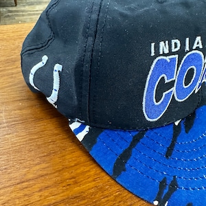 May include: A black and blue baseball cap with a white and blue camouflage pattern on the brim. The front of the cap has the words "Indiana Colts" embroidered in blue and white.