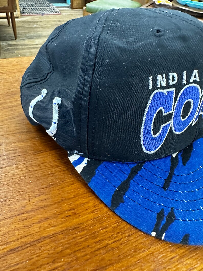 May include: A black and blue baseball cap with a white and blue camouflage pattern on the brim. The front of the cap has the words "Indiana Colts" embroidered in blue and white.