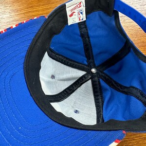 May include: Blue and white baseball cap with a red stripe. The cap has a mesh back and a black strap. The inside of the cap has a black label with the text "Ziaz" and "NBA Licensed Product".